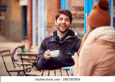 Couple On Date Sitting Outside Coffee Shop On Busy City High Street Looking At Digital Devices