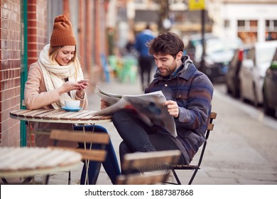 Couple On Date Sitting Outside Coffee Shop On High Street Using Mobile Phone And Reading Newspaper - Powered by Shutterstock