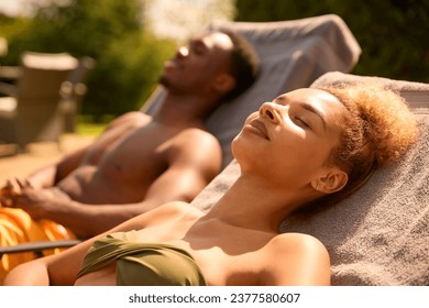 Couple On Holiday Wearing Swimming Costumes Sunbathing On Loungers By Hotel Swimming Pool