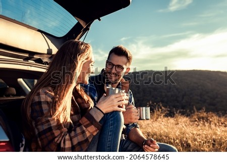 Couple on road trip sitting in trunk of a car resting and drinking coffee.