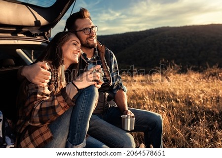 Couple on road trip sitting in trunk of a car resting and drinking coffee.