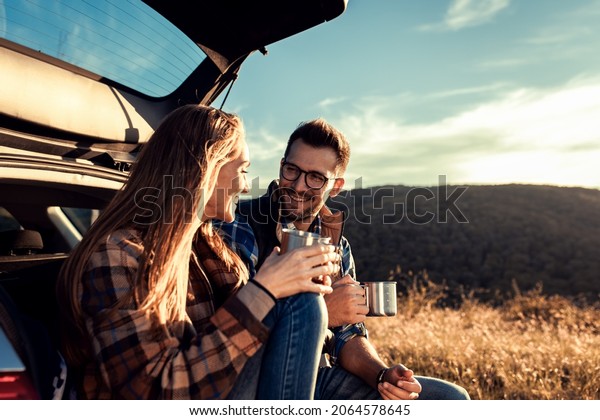 Couple on road trip sitting in trunk of a car\
resting and drinking\
coffee.