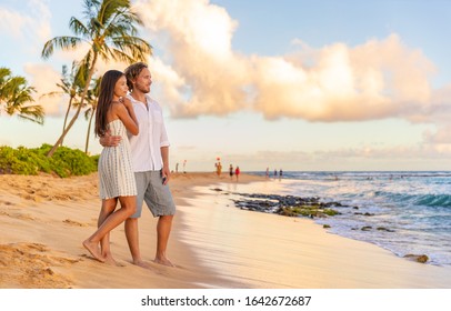 Couple On Romantic Sunset Beach Walk Relaxing During Hawaii Summer Travel Vacation On Kauai Island, USA. Asian Woman In White Dress, Man In Linen Shirt.