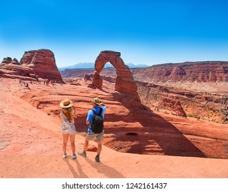 Couple On Vacation Hiking Trip. Man And Woman Standing On Top Of The Mountain Looking At Beautiful View. Delicate Arch,  Moab, Utah, Arches National Park.