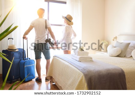 Couple on vacation with suitcases holding hands on their backs looking out a window in a bright hotel room.
