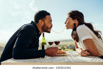 Couple On A Wine Date Sitting At A Restaurant Eating Pasta Together. Couple On A Date Spending Time Together Having Fun Holding A Strand Of Spaghetti In Their Mouth.