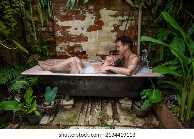 A couple are in an outdoor foam bath. She is relaxing with her feet on the edge of the bath and her head is thrown back with a blissful look on her face while he massages her shoulders. 