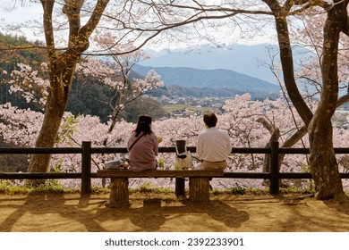 A couple picnic on a wood bench under Sakura trees, enjoying the panoramic view and Hanami (a leisure activity of admiring cherry blossoms in spring), in Takato Castle Ruins Park, Ina, Nagano, Japan - Powered by Shutterstock