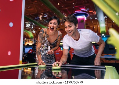 Couple Playing Air Hockey Game Holding Strikers At A Gaming Parlour. Excited Man And Woman Having Fun Playing Games At A Gaming Arcade.