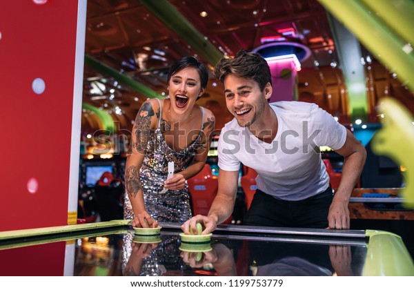 Couple playing air hockey game holding strikers\
at a gaming parlour. Excited man and woman having fun playing games\
at a gaming arcade.