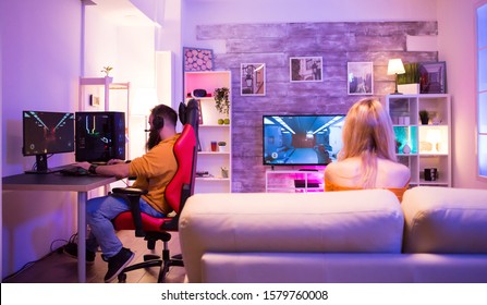 Couple Playing Different Shooter Games In Their Room With Colorful Neon Light. Man Sitting On Gaming Chair.