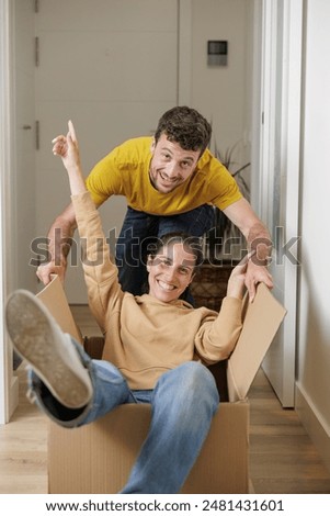 Couple playing and having fun while moving to their new house. Real estate, mortgage, rent. Woman inside a moving box, the man pushes the box down the hallway having fun.