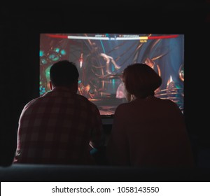 A Couple Playing Video Games In The Evening On A Big TV. Gamers Sit On The Couch At Home And Play Games On The Console. Silhouettes On The Background Of The Screen With The Game.