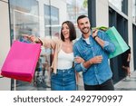 A couple points at the display of a store in the mall, deciding whether to buy an offer. A customer man and a client woman with shopping bags, smiling and looking at the shop window to purchase a gift