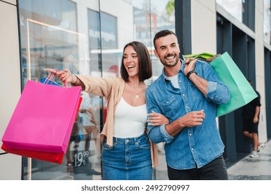 A couple points at the display of a store in the mall, deciding whether to buy an offer. A customer man and a client woman with shopping bags, smiling and looking at the shop window to purchase a gift