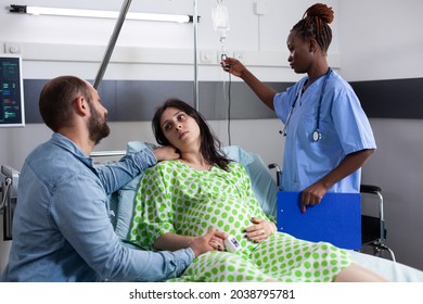 Couple With Pregnancy Waiting On Child Delivery In Hospital Ward At Clinic. African American Nurse Checking IV Drip Bag While Pregnant Woman Laying In Bed And Man Comforting About Childbirth