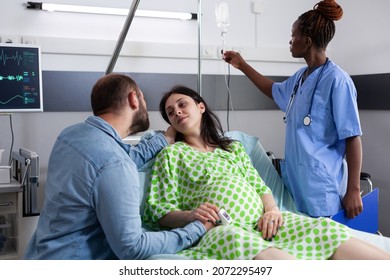 Couple With Pregnancy Waiting On Child Delivery In Hospital Ward At Clinic. African American Nurse Checking IV Drip Bag While Pregnant Woman Laying In Bed And Man Comforting About Childbirth