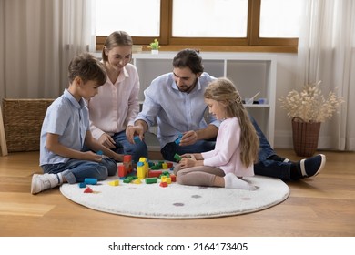 Couple And Preschool Kids, Beautiful Wellbeing Family With Children Spend Leisure, Playing Together At Home Build With Wooden Cubes Seated On Carpet In Modern Warm Nursery. Playtime, Fun, Bond Concept