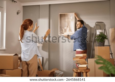 A couple is in the process of decorating their new home as one partner hangs a framed picture on the wall while the other gestures enthusiastically, surrounded by unpacked boxes and furniture.