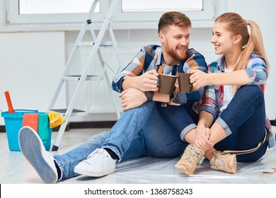 Couple Of Professional Painters Redo A Living Room, Have A Rest With A Cup Of Tea, Sitting Among Paint Tools And Accessories During The Break