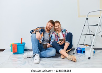 Couple Of Professional Painters Redo A Living Room, Have A Rest With A Cup Of Tea, Sitting Among Paint Tools And Accessories During The Break