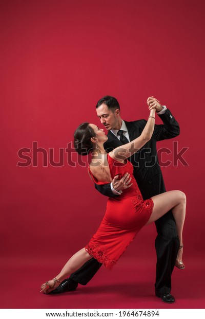 Couple of professional tango dancers\
in elegant suit and dress pose in a dancing movement on red\
background. Handsome man and woman dance looking  eye to\
eye.