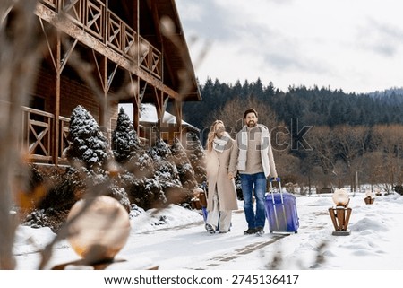 Couple pulling suitcases, walking through snow toward a wooden cabin resort for a winter vacation