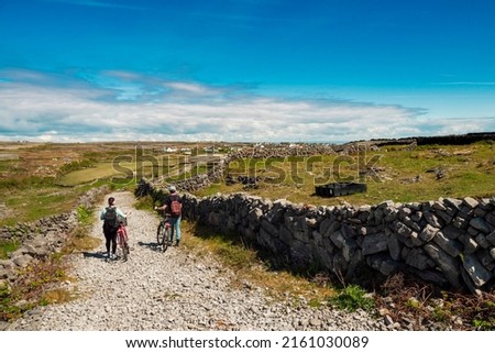 Couple pushing bikes on a rough small road. Aran island, county Galway, Ireland. Warm sunny day. Rough stone terrain. Blue cloudy sky. Popular travel area with stunning scenery. Tourism industry.