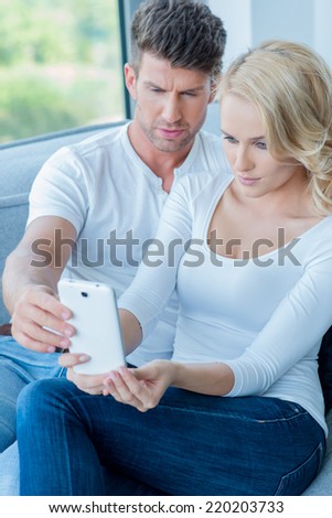 Couple reading a text message on their mobile phone with consternation frowning at the screen as they sit together on a sofa