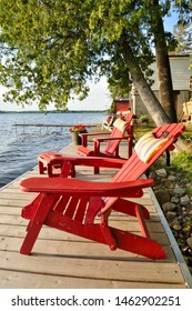 A Couple Of Red Adirondack Chairs On A Deck By The Water With A Tree And Dock In The Background