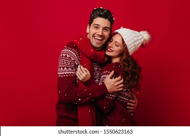 Couple In Red Christmas Sweaters Posing On Bright Background. Dark-haired Man With Friendly Smile Hugs Girl In White Knitted Hat While Snow Falls