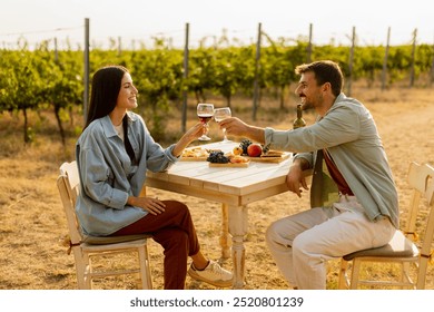 Couple relaxes at a rustic table in a sunlit vineyard, savoring wine and gourmet snacks. They share laughter and joy, surrounded by lush vines and the warmth of the evening - Powered by Shutterstock