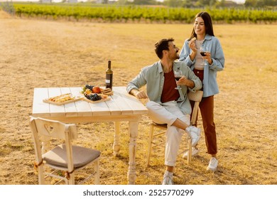 Couple relaxes at a rustic table in a sunlit vineyard, savoring wine and gourmet snacks. They share laughter and joy, surrounded by lush vines and the warmth of the evening - Powered by Shutterstock