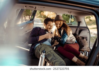 Couple relaxing in car trunk while reviewing photos on camera during road trip - Powered by Shutterstock