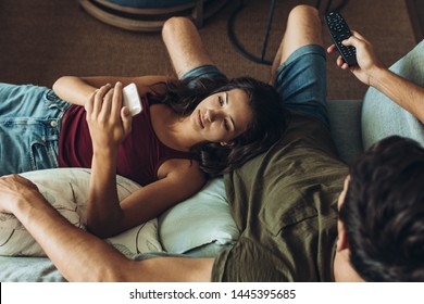 Couple Relaxing In Sofa In Front Of The TV. Woman Spending Time On Her Phone While Boyfriends Watching TV.