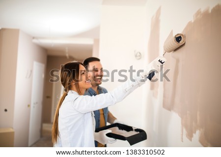 Couple renovating their apartment while man holding painting container and woman painting walls with roller in light brown color.