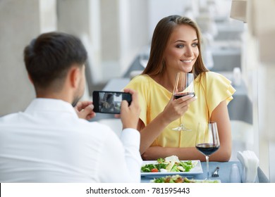 Couple In Restaurant. Man Making Photo Of Woman On Phone. Happy Smiling Young Couple Taking Photos On Mobile Phone During Romantic Dinner In Luxury Restaurant, Celebrating Holiday. High Quality Image