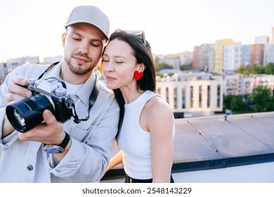 Couple reviewing photos together on rooftop. Man holding camera, woman with red heart earrings, sharing moment at sunset. Caucasian male in casual attire, lively, creative scene, photography and love. - Powered by Shutterstock