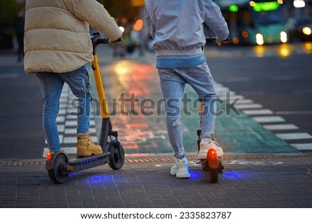 Couple rides around city at night on rent scooter in the evening. Trendy couple on electric scooter stand on crosswalk at night on city street. E-scooter or motorized vehicle for walking around.