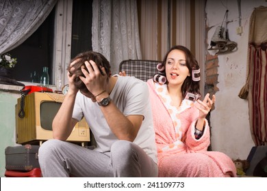 Couple In A Run Down Retro Room Sitting Arguing With The Wife In Her Dressing Gown Gesturing And Talking While The Man Covers His Ears