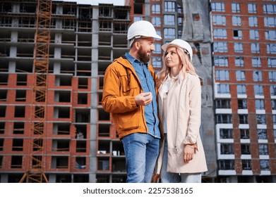 Couple in safety helmets standing outside apartment building under construction. Man and woman discussing building plan outdoors at construction site.