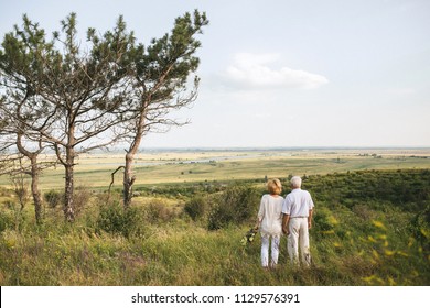 A Couple Of Senior Adults In The White Linen Dress Is Standing With Backs Outdoors, In The Hands A Bouquet Of Flowers, Three Coniferous Trees, Background Of Beautiful Landscape And Sky. Horizontal