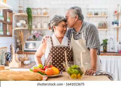 Couple Senior Asian Elder Happy Living In Home Kitchen. Grandfather Cooking Salad Dish With Grandmother With Happiness, Kissing Enjoy Retirement Life Together. Older People Relationship And Activity.