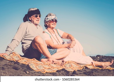 Couple Of Senior Caucasian Hippy Style And Life Rest On The Rocky Beach In Tenerife. Colors And Alternative Lifestyle For Totally Freedom Concept Near The Ocean.