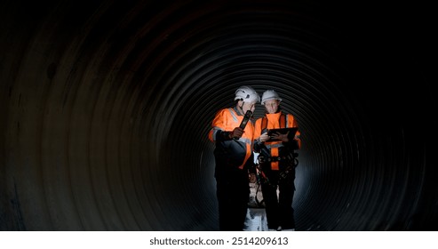 A couple of senior civil engineers are collaborating in the tunnel of a sewage drain, using a digital tablet to inspect the construction of a huge water pipe on a new highway road project. - Powered by Shutterstock