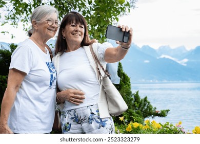 Couple of senior females friends walking outdoors along Lake Geneva while take a selfie with smartphone. Smiling elderly women  enjoying freedom and vacation or retirement - Powered by Shutterstock