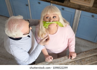 Couple Of Senior People Having Fun While Making Lunch. Charming Old Woman Being Silly Putting Lettuce On Her Upper Lip So It Looks Like Mustache.