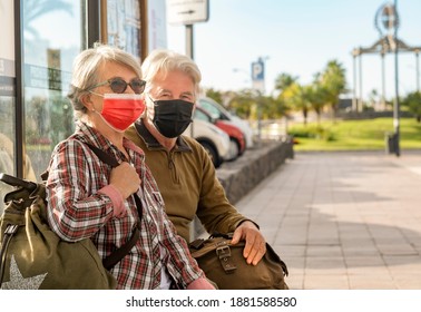 Couple Of Senior Travelers Sitting On The Bench While Waiting For The Bus Wearing A Surgical Mask Due To The Coronavirus. Active Retired People Enjoying Travel And Freedom