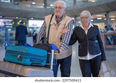 Couple of senior travelers with trolley and suitcases pushing luggage trolley while walking through airport area to reach departure gate. Old senior man and woman leaving for vacation - Powered by Shutterstock