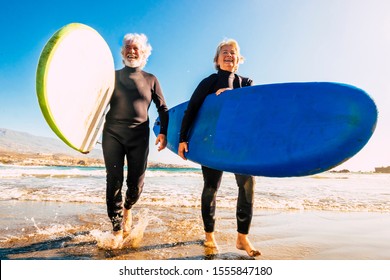 couple of seniors at the beach with black wetsuits holding a surftable ready to go surfing a the beach - active mature and retired people doing happy activity together in their vacations or freetime